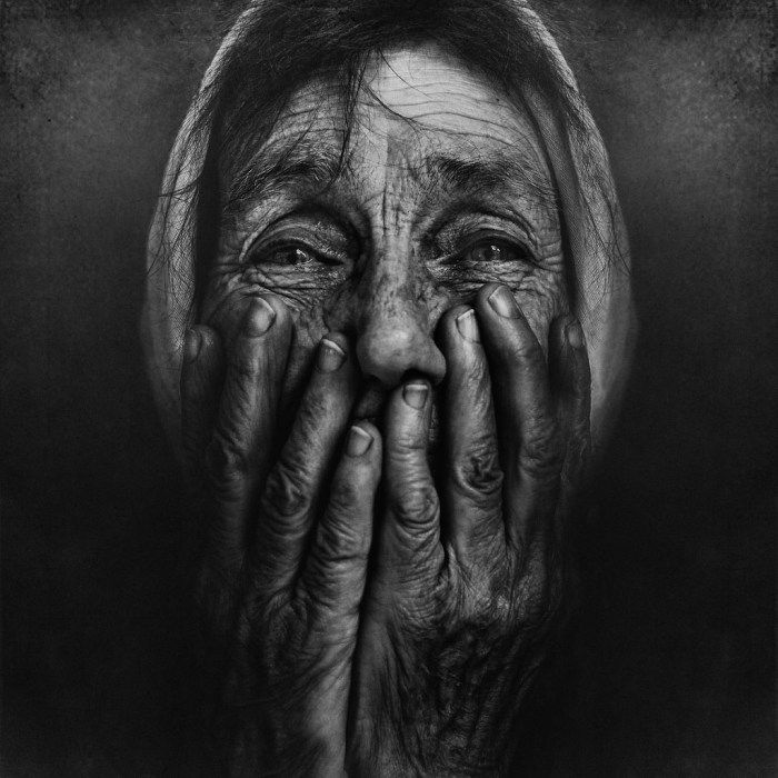 Dramatic monochrome fine art photography of a person covering their face entirely with wrinkled weathered hands.