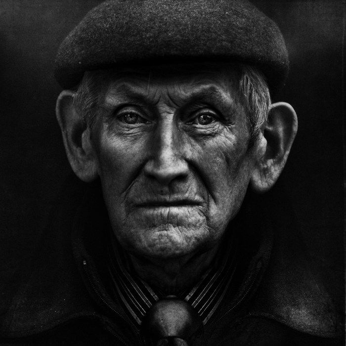 Sharp focus monochrome portrait of an old man wearing a flat cap staring directly into the camera lens.