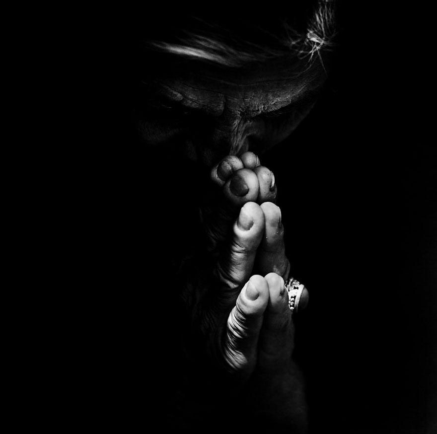 Intense monochrome portrait of a homeless person with weathered hands clasped near face in dark low key lighting.
